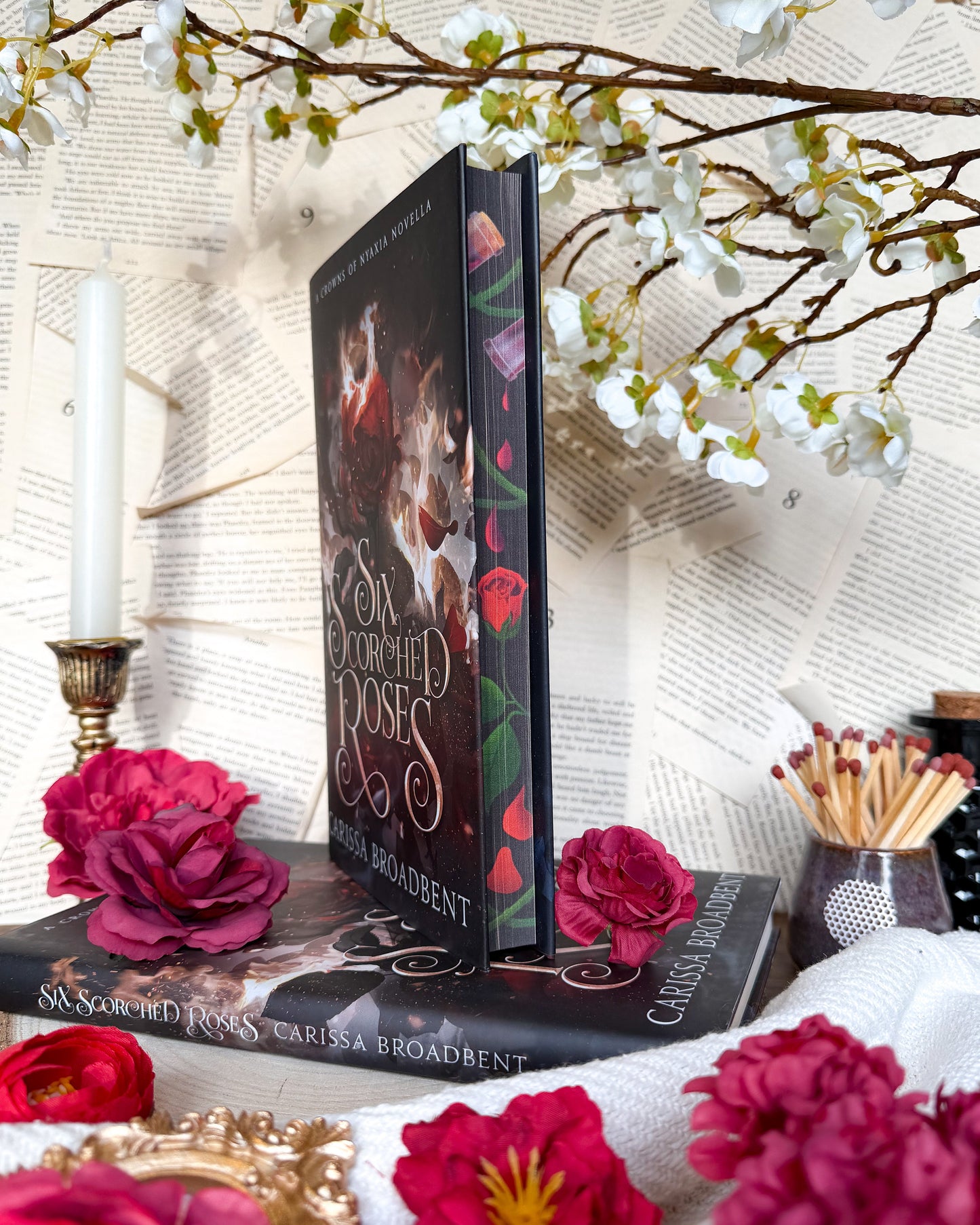 Book titled 'Six Scorched Roses' by Carissa Broadbent on a decorative surface with flowers and candles.