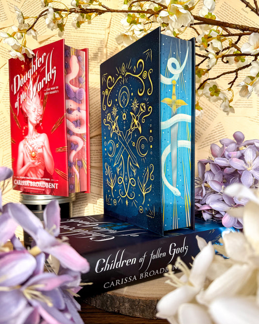 A book titled ''Children of Fallen Gods' by Carissa Broadbent, surrounded by flowers, with a book titled 'Daughter of No Worlds' visible in the background.