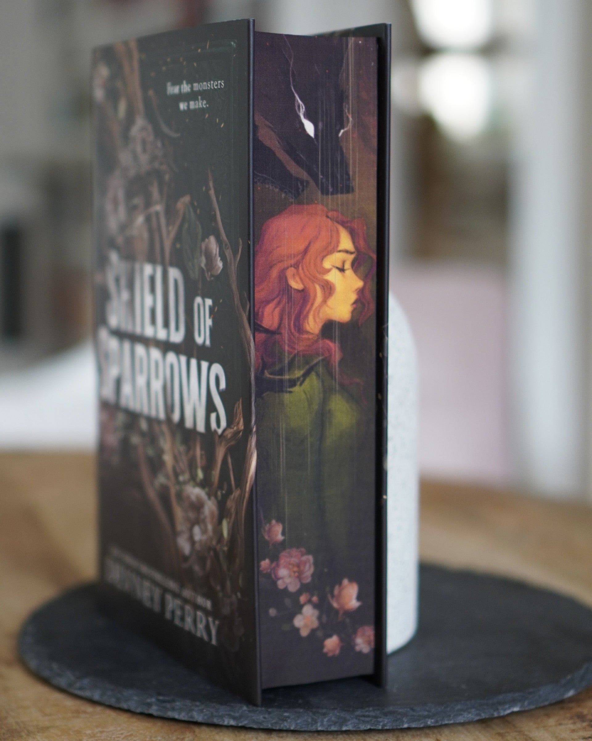Book titled 'Shield of Sparrows' with a woman's face on the cover, displayed on a stand.
